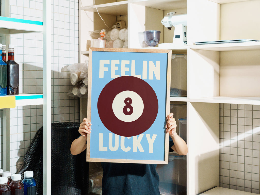 A person in their home holds up a framed art print featuring a light blue background, a brown billiard 8-ball, and the words 'FEELIN LUCKY' in cream. The background shows white tiled walls and shelves with various store items.