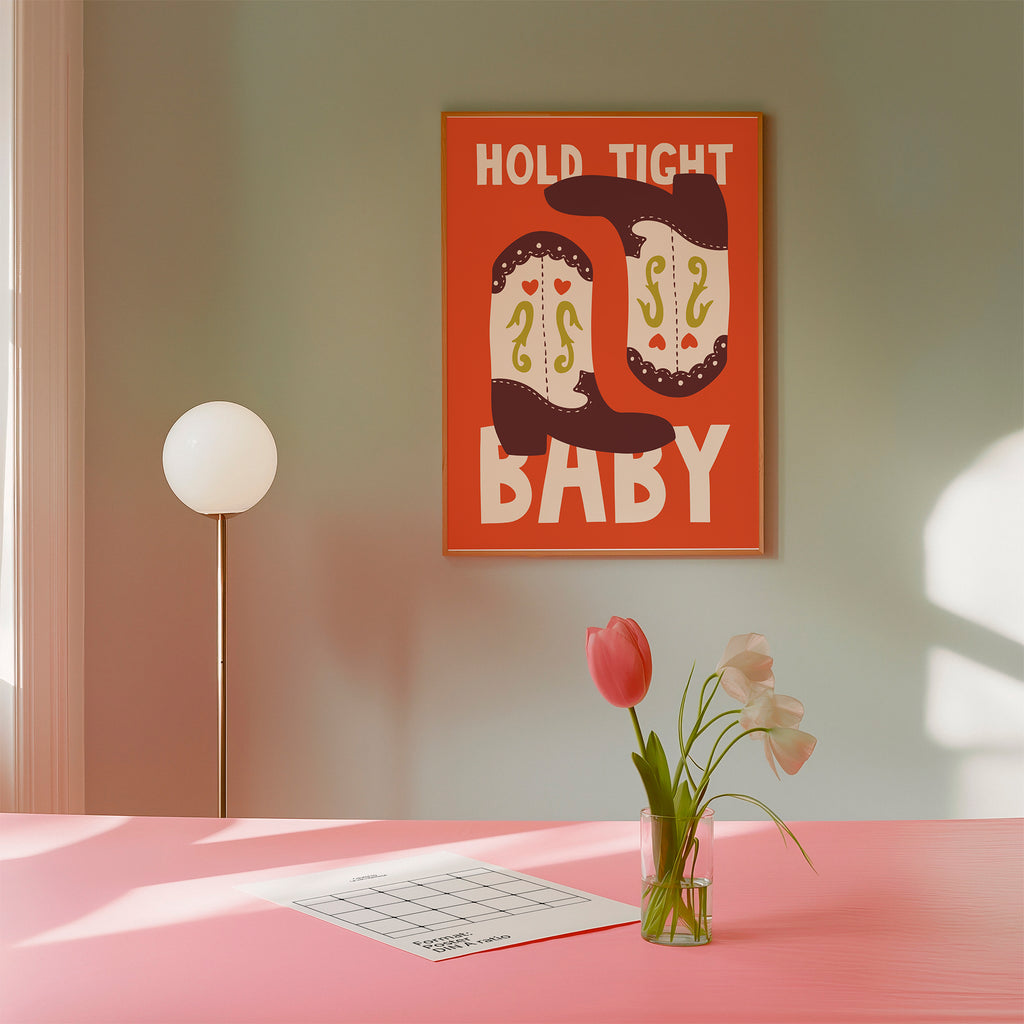 A framed wall art print with the words “HOLD TIGHT BABY” and two cowboy boots hangs on a muted green wall above a pink table. A floor lamp stands to the left, and a small vase with tulips and a paper tray sit on the table in the foreground.