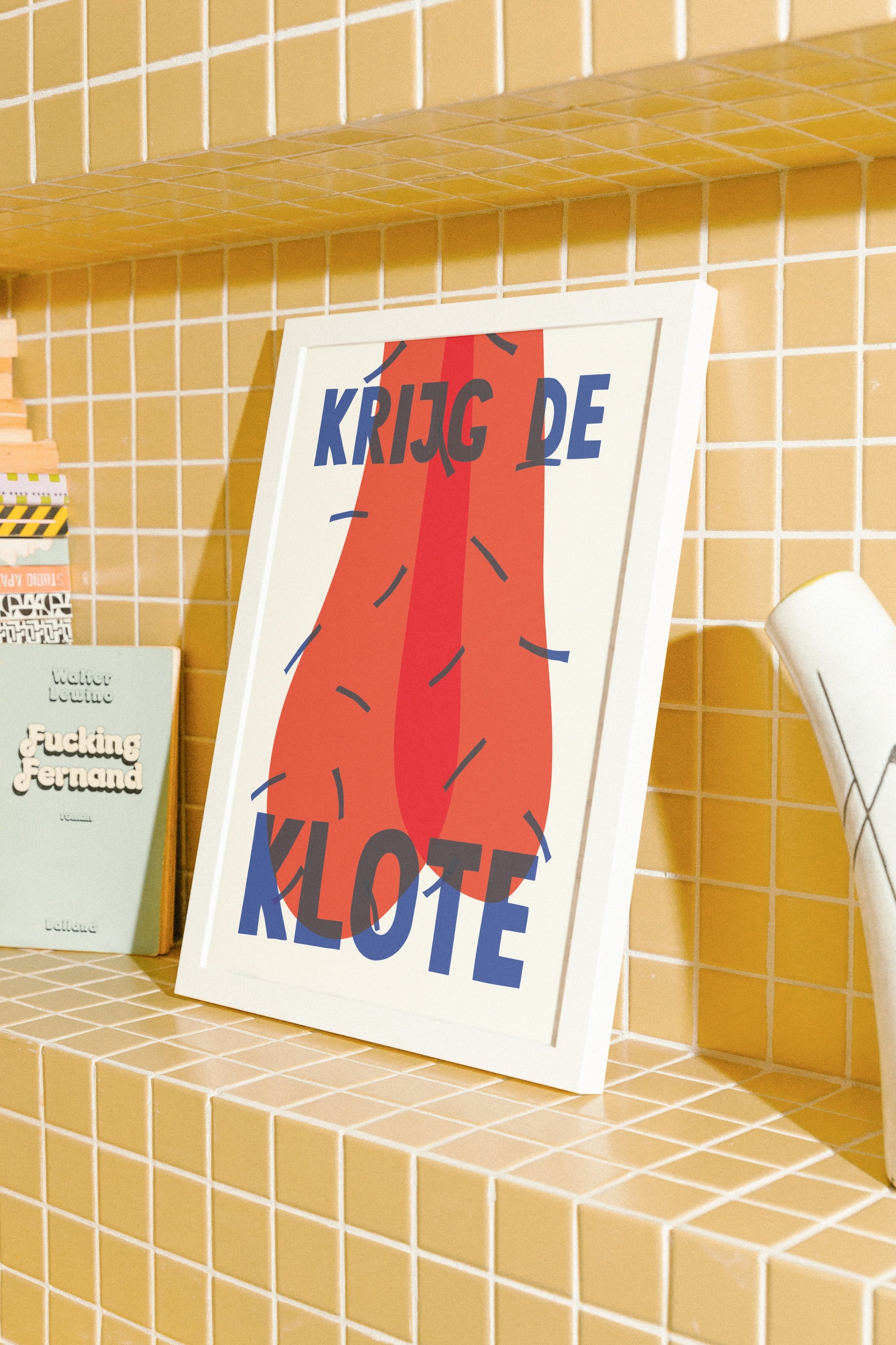 A framed poster with the Dutch text 'KRIJG DE KLOTE' and a large red abstract shape, leaning on a yellow tiled shelf next to a small book.