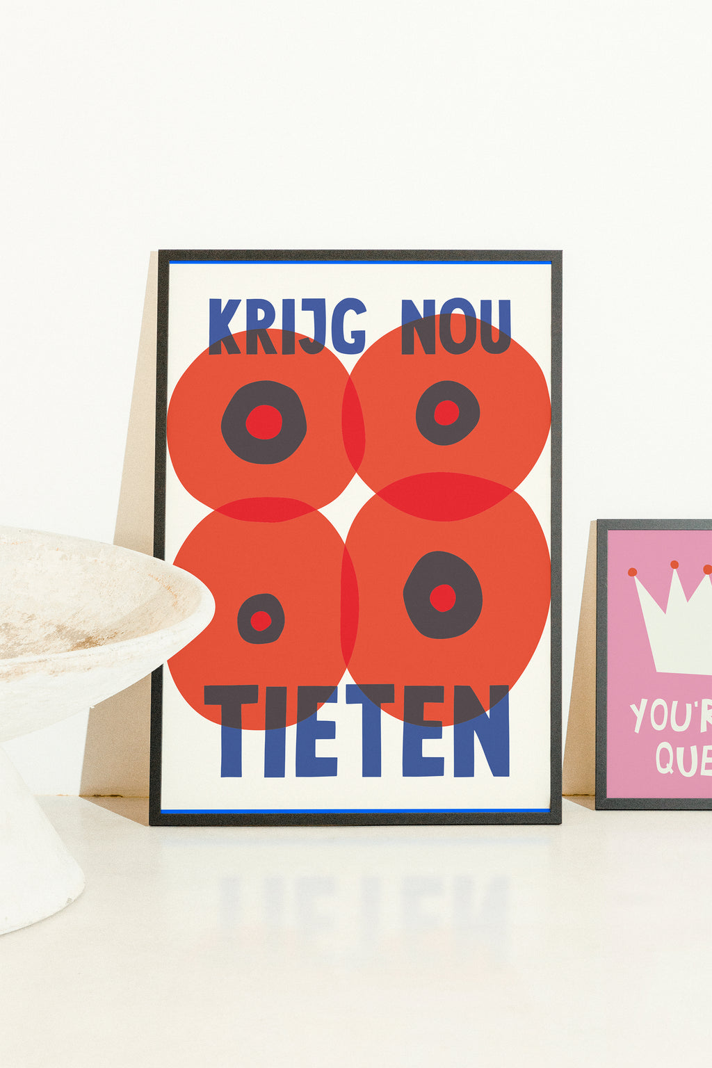 A framed graphic art print featuring four red poppy-like circles with black centers and blue text reading 'KRIJG NOU TIETEN' on a white background, styled on a white surface next to a partial pink framed print.
