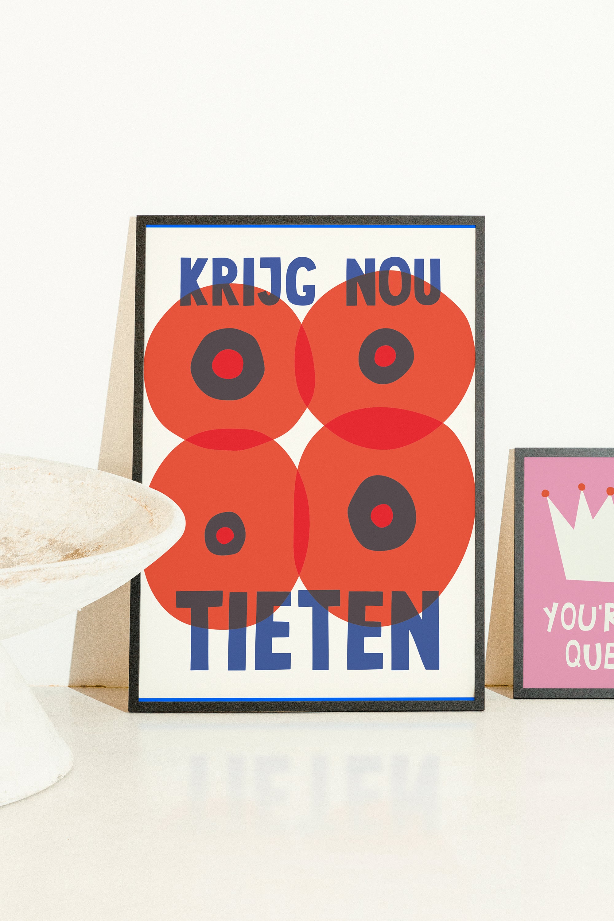 A framed graphic art print featuring four red poppy-like circles with black centers and blue text reading 'KRIJG NOU TIETEN' on a white background, styled on a white surface next to a partial pink framed print.